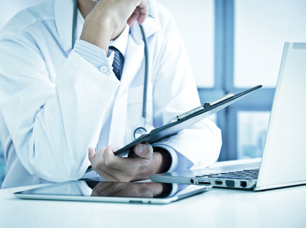 Picture of a male Physician sitting at a desk with a laptop open and holding a clipboard. There is a tablet next to the laptop.